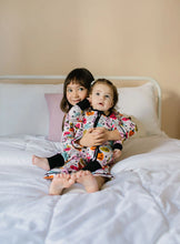 Load image into Gallery viewer, Two children sitting together on a bed wearing matching Halloween-themed pajamas with a colorful pumpkin and spider print on a white background.
