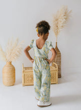 Load image into Gallery viewer, Child wearing a floral jumpsuit standing in front of decorative vases and a woven basket.
