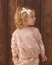 Load image into Gallery viewer, A young child with blonde curly hair wearing a dusty pink long-sleeve sweatshirt and matching pants, decorated with a white ghost pattern. The child is wearing a white bow in their hair and standing against a dark brown wooden plank background.
