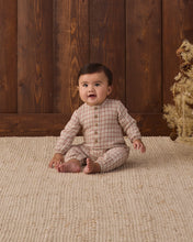 Load image into Gallery viewer, Baby sitting barefoot on a cream rug wearing a Christmas plaid jumpsuit beside a small decorative tree.
