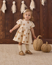 Load image into Gallery viewer, A toddler girl stands in a styled indoor setting wearing a cream dress with brown pumpkin prints, white socks, and tan shoes. The background features a dark wood plank wall with hanging fabric tassels and two woven decorative pumpkins on a textured rug.
