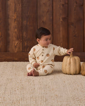 Load image into Gallery viewer, A baby sitting on a textured cream rug in front of a dark wood plank wall, wearing a cream long-sleeve pajama top with brown pumpkin prints. The baby is touching a small natural pumpkin prop.
