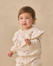 Load image into Gallery viewer, A toddler wearing a cream-colored ruffled long-sleeve top and matching pants with small dark embroidered dots, holding a small bunch of dried flowers against a warm beige studio background.
