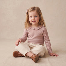 Load image into Gallery viewer, A toddler girl sitting cross-legged on a textured beige studio backdrop, wearing a dusty pink long-sleeve top with a ruffled collar, cream pants, and brown shoes.
