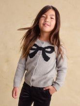 Load image into Gallery viewer, A young girl with long brown hair wearing a light gray knit cardigan with a large black bow motif on the chest, paired with black pants, standing against a textured beige background.

