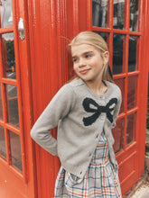 Load image into Gallery viewer, A young girl with blonde hair stands in front of a red telephone booth wearing a gray knit cardigan with a black bow motif and a plaid skirt.
