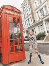 Load image into Gallery viewer, A young girl in a grey long-sleeve top, plaid skirt, and blue knee-high socks stands next to a classic red London telephone booth on a city street with white townhouse buildings in the background.
