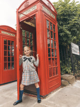 Load image into Gallery viewer, A young girl in a grey dress with a black bow and plaid skirt stands smiling inside an open red telephone booth, with another red telephone booth and green foliage in the background.
