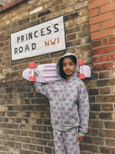 Load image into Gallery viewer, A child wearing a light gray zip-up hoodie and matching pants with a purple bow pattern, holding a pink cruiser skateboard against a brick wall with a 'Princess Road NW1' sign.
