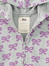 Load image into Gallery viewer, Close-up of a light gray zip-up hoodie with an allover purple bow pattern, featuring a cream-colored interior neck label with the brand name 'tea'.

