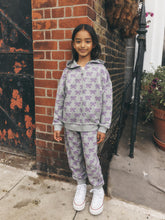 Load image into Gallery viewer, A young girl stands on a sidewalk in front of a red brick wall, wearing a light gray zip-up hoodie and matching jogger pants with a purple bow pattern, paired with white sneakers.

