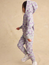 Load image into Gallery viewer, A child wearing a light gray two-piece tracksuit with a purple bow pattern, featuring a hooded sweatshirt and matching jogger pants, posed against a textured beige background.
