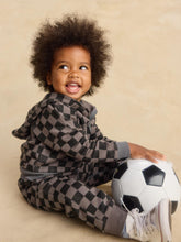 Load image into Gallery viewer, A smiling toddler with curly dark hair sits on a beige studio floor wearing a matching black and gray checkered hooded sweatshirt and pants, with a black-and-white soccer ball beside them.
