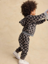 Load image into Gallery viewer, A toddler wearing a matching gray and black checkered hoodie and jogger pants set, paired with white sneakers, captured from a side-back angle against a warm beige studio backdrop.
