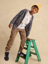 Load image into Gallery viewer, A child model wearing a gray and black checkered zip-up hoodie, a white t-shirt with a beer graphic, brown pants, and a yellow beanie, leaning on a green wooden step stool against a textured beige background.
