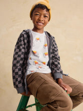 Load image into Gallery viewer, A young boy wearing a yellow knit beanie, a gray and black checkered zip-up hoodie over a white graphic t-shirt, and brown pants, sitting on a green stool against a textured beige wall.
