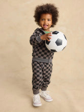 Load image into Gallery viewer, A toddler with curly dark hair stands against a beige studio backdrop, wearing a dark gray and black checkerboard sweatshirt and matching jogger pants, holding a black-and-white soccer ball.
