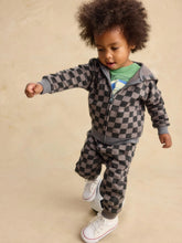 Load image into Gallery viewer, A toddler with curly dark hair wearing a gray and black checkered zip-up hoodie and matching checkered pants over a green t-shirt, standing on a white stool against a beige background.

