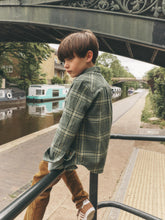 Load image into Gallery viewer, A young boy wearing a green and yellow plaid flannel shirt and brown corduroy trousers stands on a canal-side walkway under a bridge.
