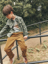 Load image into Gallery viewer, A young boy wearing a green plaid flannel shirt over a graphic t-shirt, brown corduroy pants, and brown sneakers, sitting on a black metal railing outdoors with trees in the background.
