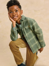 Load image into Gallery viewer, A young boy crouching in a studio setting, wearing a sage green plaid button-up shirt over a light blue undershirt and tan corduroy pants, against a warm beige textured background.
