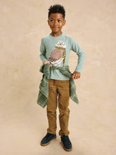 Load image into Gallery viewer, A young boy modeling a sage green long-sleeve t-shirt with a large owl graphic, styled with brown pants, navy shoes, and a green plaid shirt tied around his waist, set against a warm beige textured backdrop.
