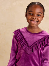 Load image into Gallery viewer, A young girl smiling at the camera, wearing a purple velvet long-sleeve top with a ruffled V-neckline, set against a warm beige textured background.
