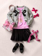 Load image into Gallery viewer, A styled flat-lay outfit for a child featuring a light gray knit cardigan with black bow details, a pink t-shirt with a cake graphic, a black pleated skirt, black shoes, a pink ribbon, and a floral gift bag on a textured light beige fabric background.
