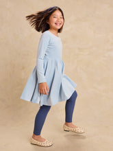 Load image into Gallery viewer, A young girl smiling and twirling while wearing a light blue long-sleeve dress, navy blue leggings, and beige polka-dot flats against a textured beige studio backdrop.
