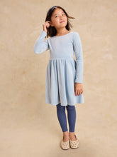 Load image into Gallery viewer, A young girl modeling a light blue long-sleeve dress with a flared skirt, paired with navy blue leggings and beige shoes, standing against a textured beige studio backdrop.
