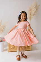 Load image into Gallery viewer, A young girl with a blue bow in her hair is wearing a short-sleeve coral pink dress with a white polka dot pattern, twilly lace trim, and a flared skirt. She is standing in front of a light-colored wall with pampas grass and a wicker chest, holding out her dress to show its volume.

