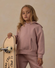 Load image into Gallery viewer, A young girl with blonde hair stands holding a skateboard, wearing a matching dusty pink hoodie and sweatpants set against a warm beige background.
