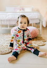 Load image into Gallery viewer, A baby sitting on a woven rug in a bedroom, wearing a white one-piece pajama with a colorful Halloween print of pumpkins, ghosts, and spiders.
