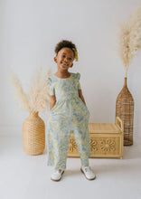 Load image into Gallery viewer, A young child wearing a light blue and yellow floral print jumpsuit stands in a studio setting with decorative woven baskets and pampas grass.
