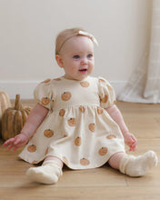 Load image into Gallery viewer, A baby sitting on a wooden floor wearing a cream-colored dress with a pumpkin print, a matching bow headband, and cream booties.

