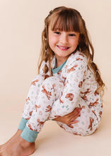 Load image into Gallery viewer, A young girl with long light brown hair sits cross-legged against a solid beige background, wearing a white long-sleeve pajama set with a brown and green woodland animal print and light blue cuffs.
