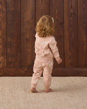 Load image into Gallery viewer, A toddler with curly blonde hair stands barefoot from the back, wearing a light pink two-piece pajama set with a white cloud pattern, set against a dark brown wooden plank wall and a beige woven rug.
