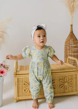 Load image into Gallery viewer, A baby girl wearing a light sage green and yellow floral print romper with a white bow headband, standing barefoot in front of a wicker chest.
