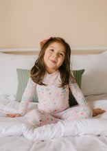 Load image into Gallery viewer, Child sitting cross-legged on white bedding in Sugar Plum pajamas with a pastel holiday print.
