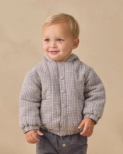 Load image into Gallery viewer, A young toddler with blonde hair wearing a brown and white checkered hooded puffer jacket and dark gray pants, standing against a solid beige background.
