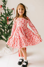 Load image into Gallery viewer, Child holding out a pale pink dress with red bow-and-stripe print, long sleeves, and a wide ruffled hem.
