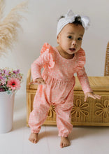 Load image into Gallery viewer, A baby wearing a pink long-sleeve romper with white polka dots and a matching white bow headband, standing barefoot in front of a wicker chest.
