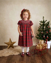 Load image into Gallery viewer, A young girl stands in a studio setting wearing a red and black plaid dress with white trim, white tights, and dark red shoes. She is posed next to a decorated Christmas tree, a red sleigh with wrapped gifts, and a gold star on a wood floor against a textured beige backdrop.
