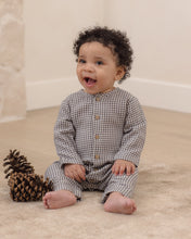 Load image into Gallery viewer, A baby with curly dark hair sits on a beige carpet next to a pinecone, wearing a long-sleeve button-front romper in a brown and beige gingham check pattern.
