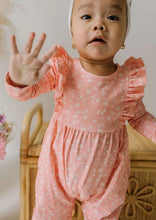 Load image into Gallery viewer, A baby wearing a pink long-sleeve romper with a white floral print, ruffled shoulders, and a gathered elastic waist, standing indoors and waving with one hand.
