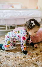 Load image into Gallery viewer, A toddler girl crawling on a woven jute rug in a bedroom, wearing a white long-sleeve pajama top and matching pants covered in colorful Halloween-themed graphics like pumpkins, ghosts, and stars. She has a white bow in her hair.
