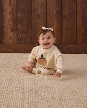 Load image into Gallery viewer, A baby wearing a cream-colored long-sleeve top with a pumpkin graphic and a white bow headband, sitting on a woven beige rug in front of a dark wood plank wall.
