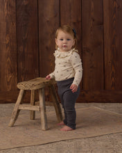 Load image into Gallery viewer, A toddler stands barefoot next to a rustic wooden stool in front of a dark wood plank wall. The child is wearing a cream long-sleeve ruffled blouse with small dark motifs and dark gray ribbed pants.
