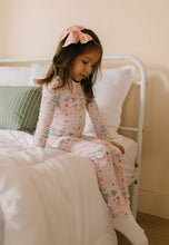 Load image into Gallery viewer, Child with a pink hair bow sitting on the edge of a bed in pale pink Sugar Plum pajamas and white socks.
