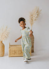 Load image into Gallery viewer, A young child wearing a light blue and yellow floral print jumpsuit stands in a bright studio setting with decorative pampas grass and a woven rattan chest.
